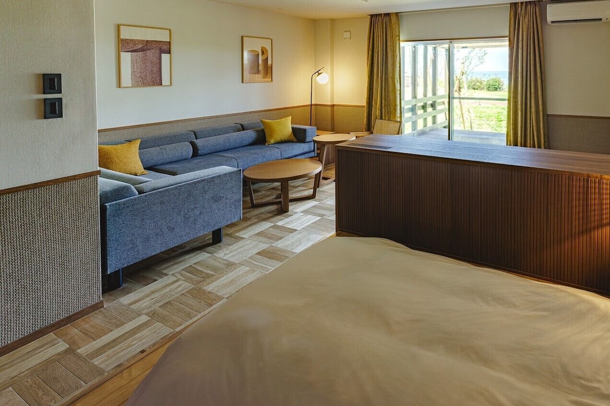 Quarto de hotel com cama, sofá, mesa pequena, escrivaninha e vista para o exterior.