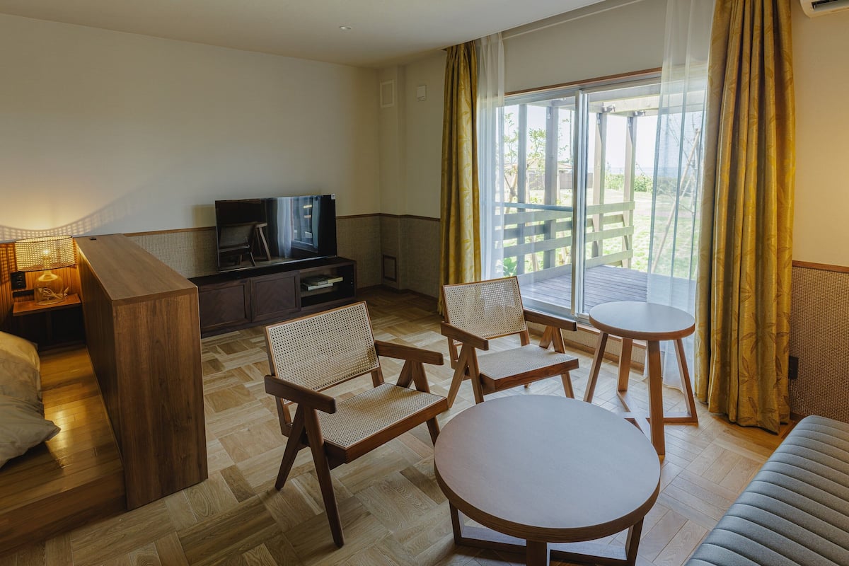 Quarto de hotel com televisão, mobiliário de madeira, varanda e vista, e uma luminária.