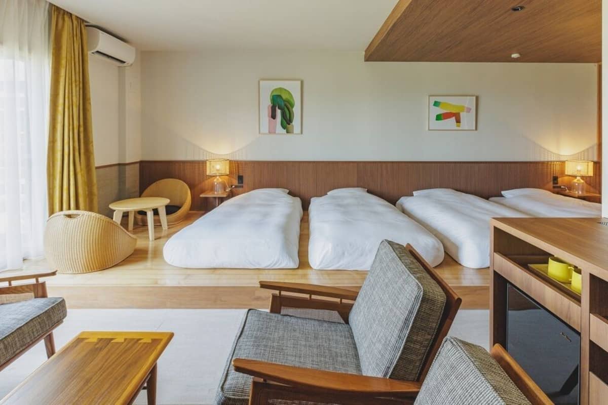 Quarto de hotel com sofá, duas camas de solteiro, uma mesa pequena, uma cadeira e televisão.
