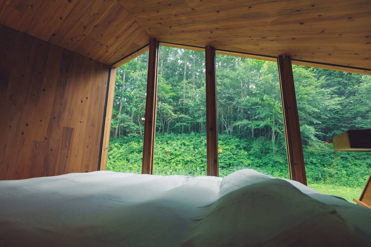 Una habitación de cabaña de madera con cama, ventanas amplias y vista a un bosque.