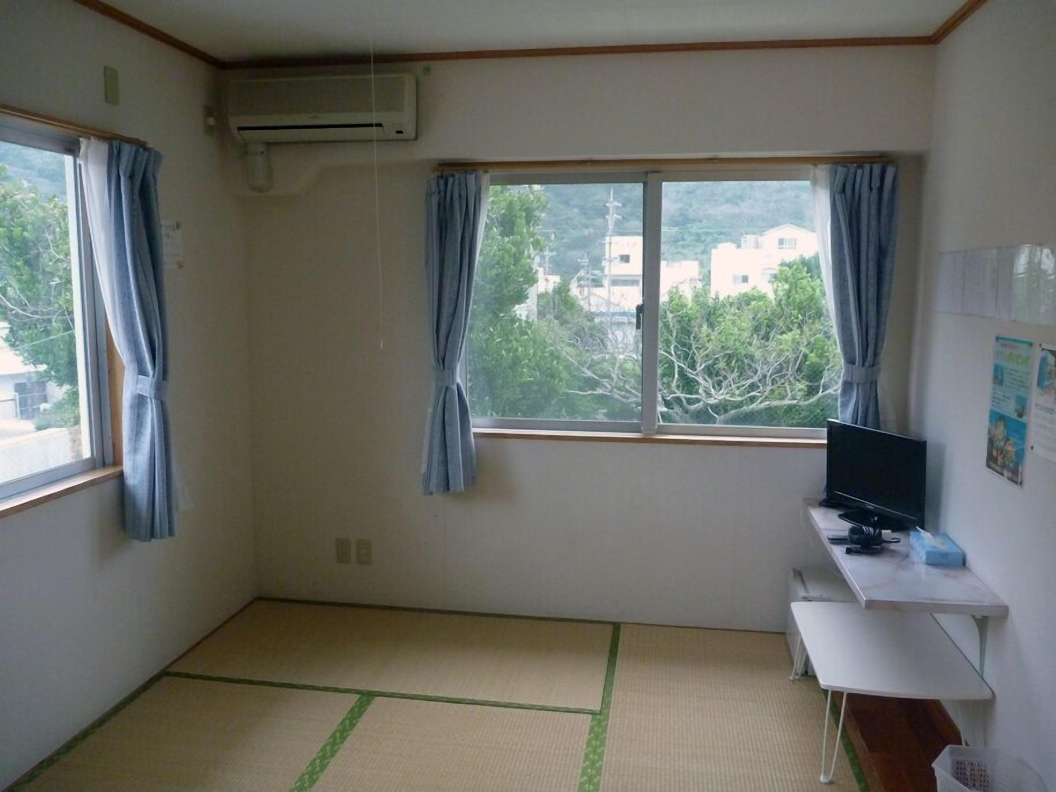 Een kamer met tatami vloerbedekking, een bureau met een flatscreen televisie en twee ramen met gordijnen.