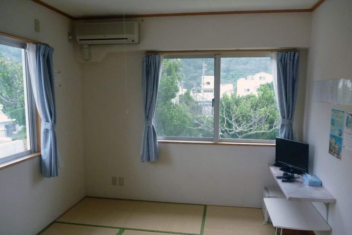 Quarto com piso de esteira de tatame, uma escrivaninha com televisão de tela plana e duas janelas com cortinas.