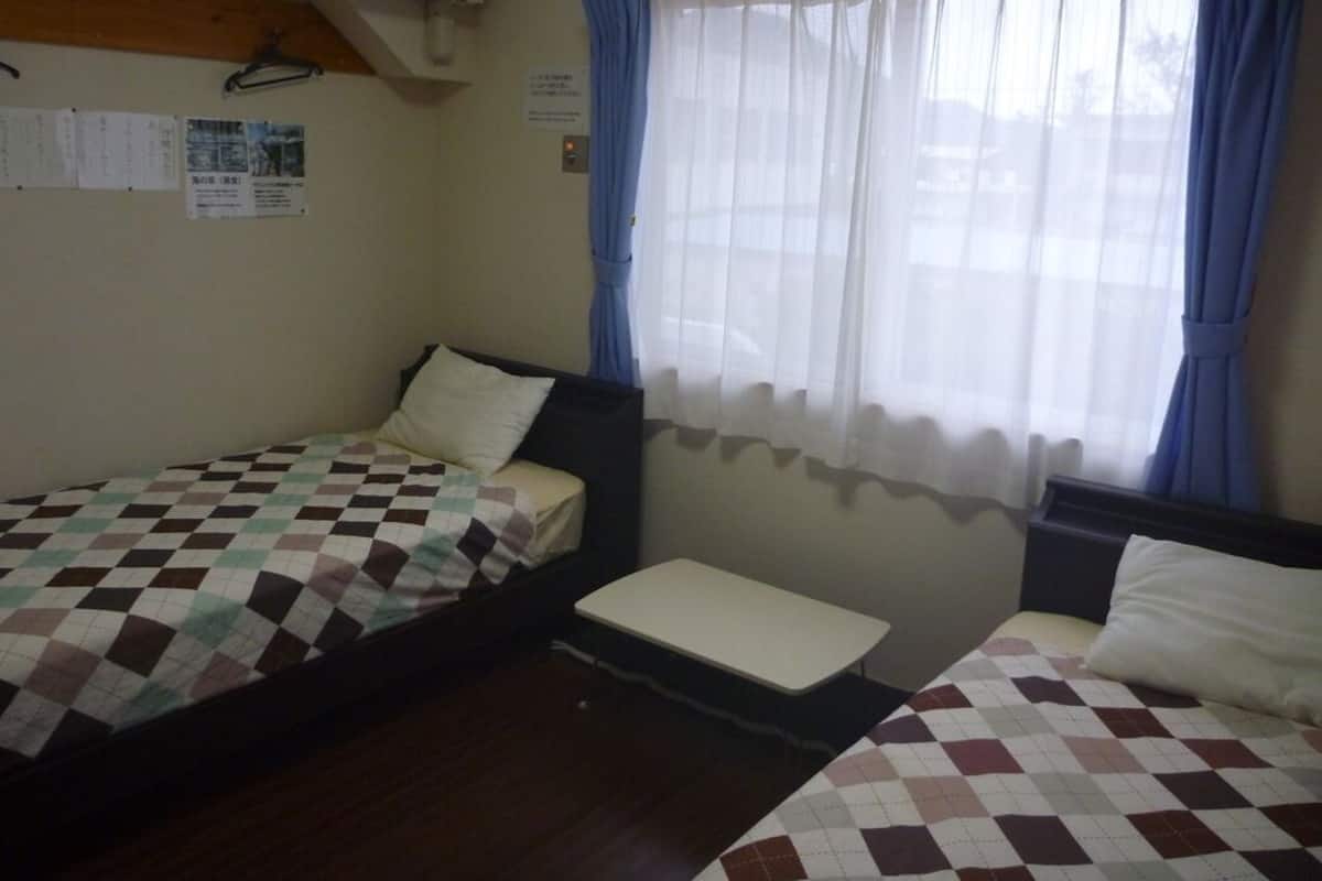 Quarto com duas camas, uma mesa pequena e uma janela com cortinas.