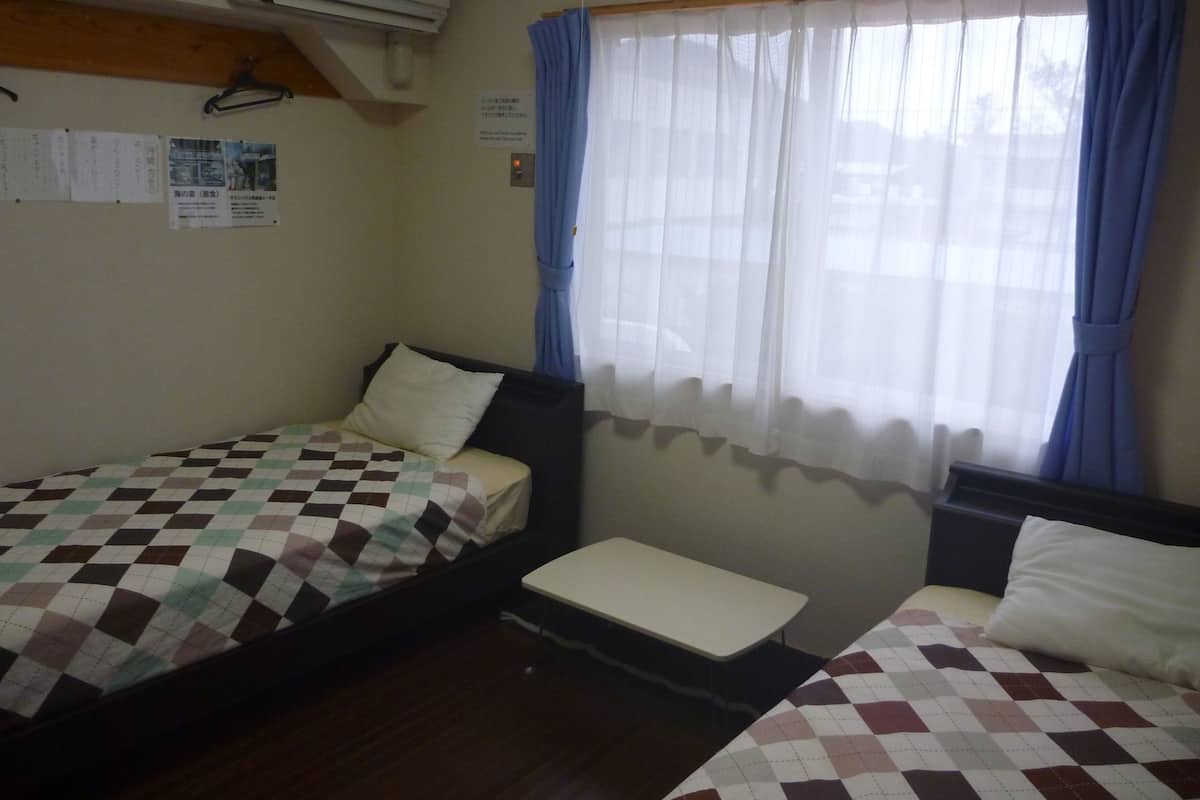 Quarto com duas camas, uma mesinha e uma janela com cortinas.