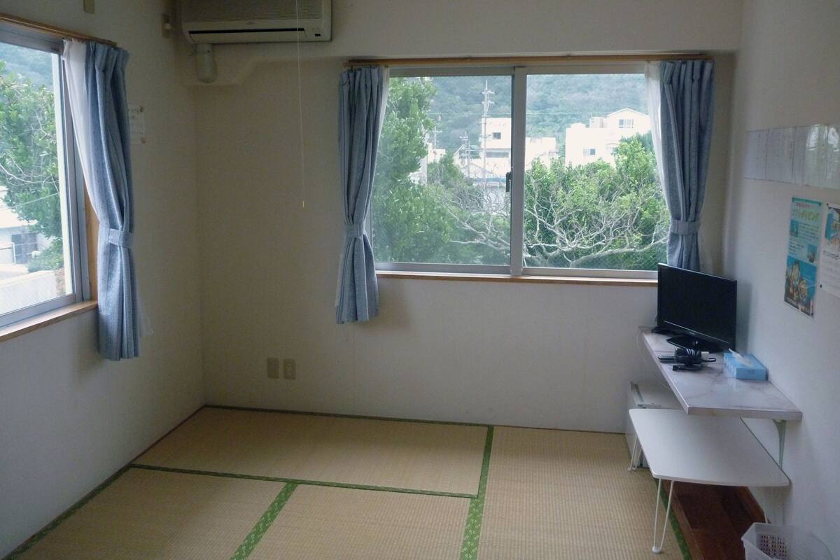 Um quarto com tatame, uma escrivaninha com computador e uma janela com cortinas.