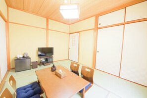 Ein Zimmer mit Tatami-Böden, einem Holztisch, Stühlen, einem Fernseher, einem kleinen Kühlschrank und einer Schiebetür.