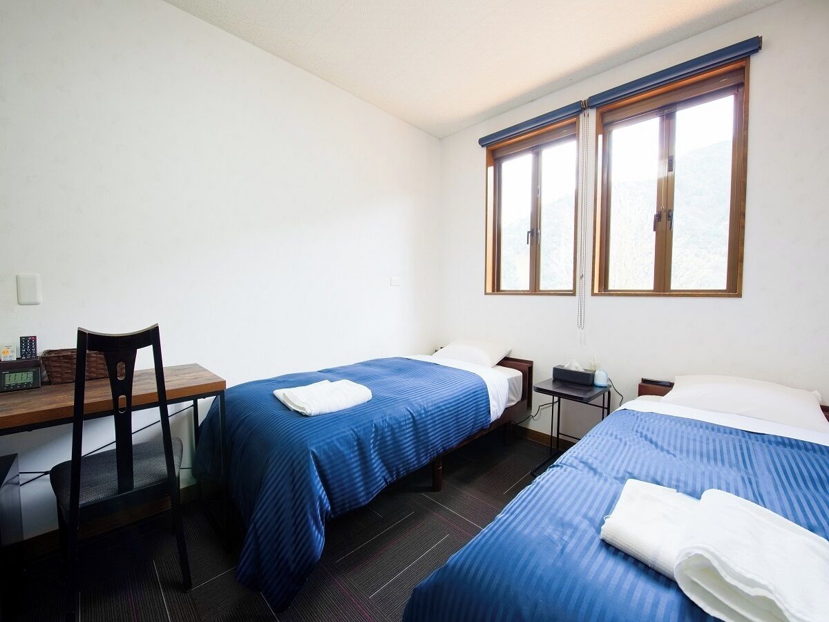A room with two beds, a desk, a chair, and a window with blinds.
