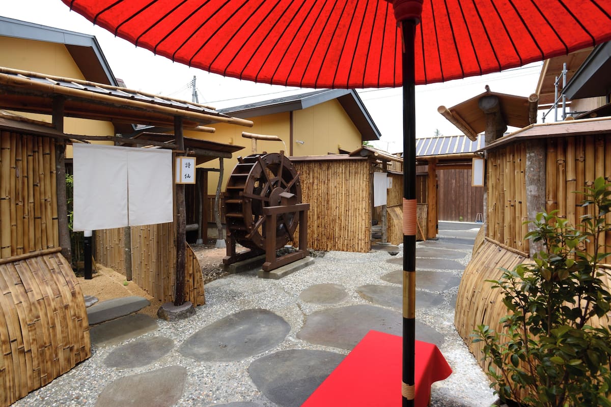 Un patio japonés tradicional con una rueda hidráulica, vallas de bambú y un paraguas rojo.