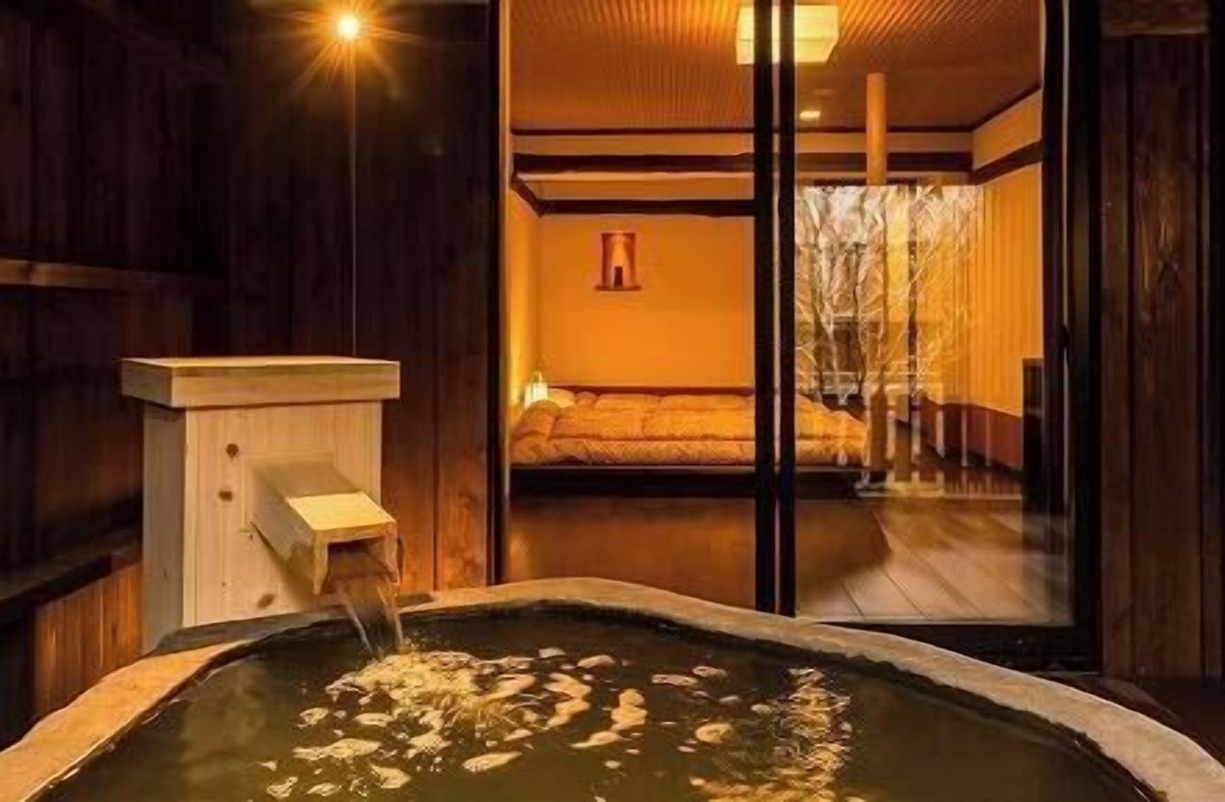 Un onsen japonés tradicional con una bañera de madera, una estructura de madera con un caño y vista a un cuarto con cama y pared de madera.