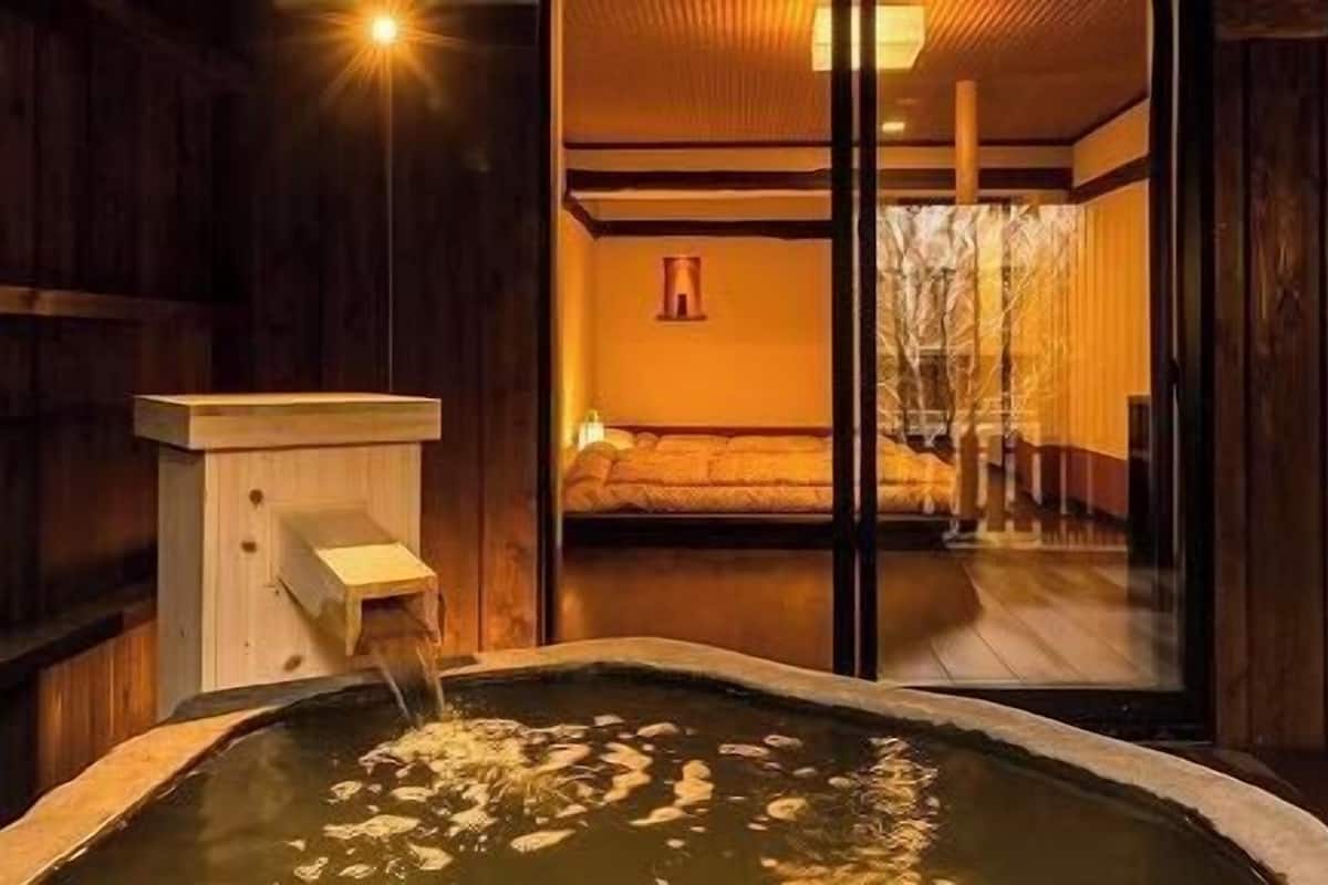 Un onsen japonés tradicional con una bañera de madera, una estructura de madera con un caño y vista a un cuarto con cama y pared de madera.