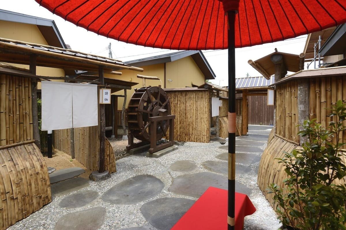 Un entorno tradicional japonés con una rueda hidráulica, vallas de bambú y un paraguas rojo.