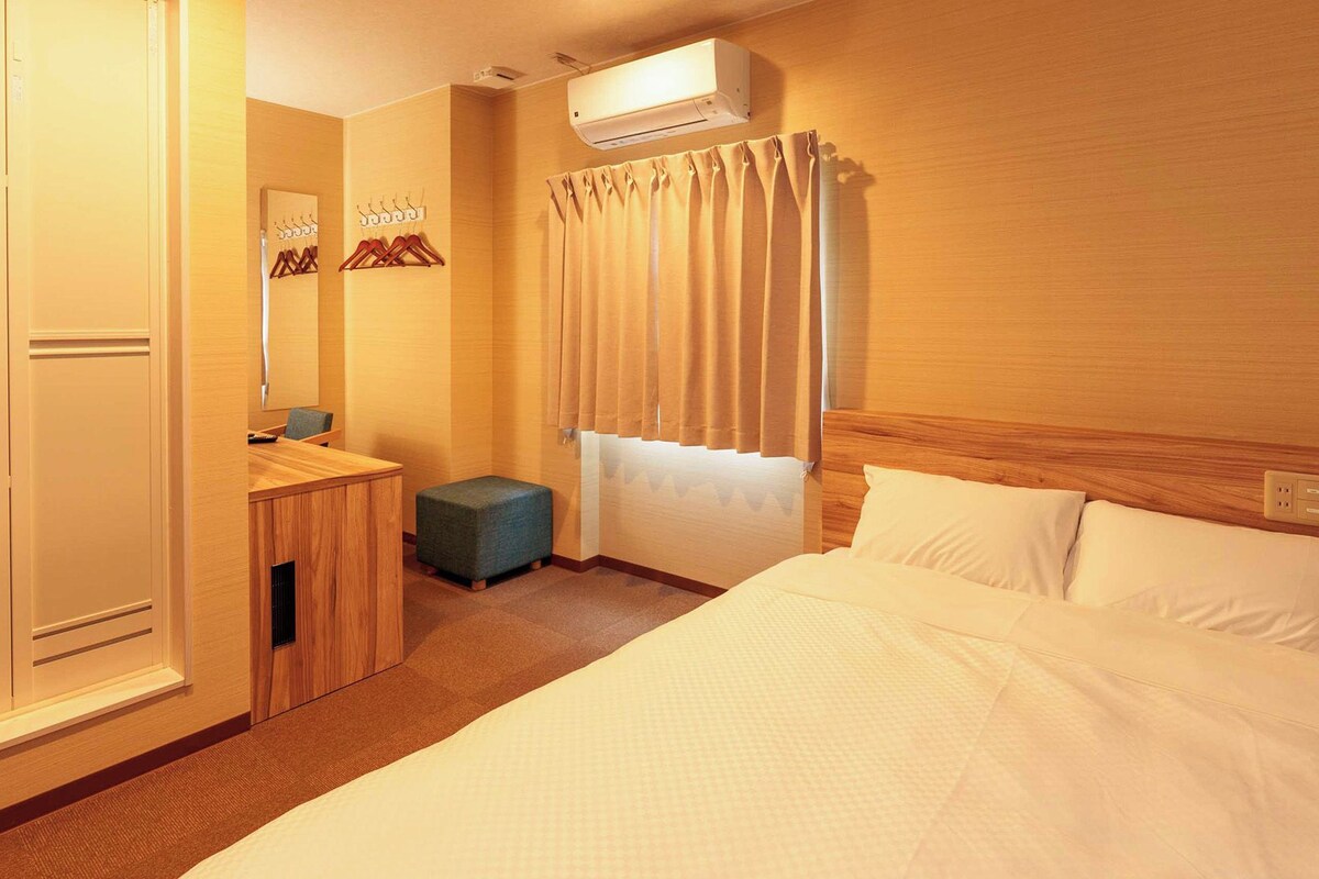 Una habitación de hotel con cama, escritorio y silla, un taburete pequeño, espejo, ventana con cortinas y equipo de aire acondicionado.