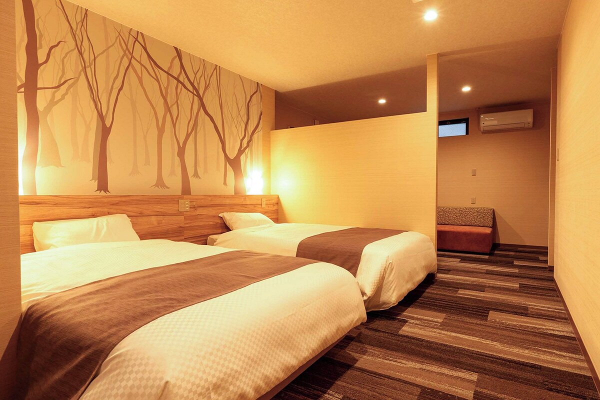 Habitación de hotel con dos camas, un televisor montado en la pared y un banco.