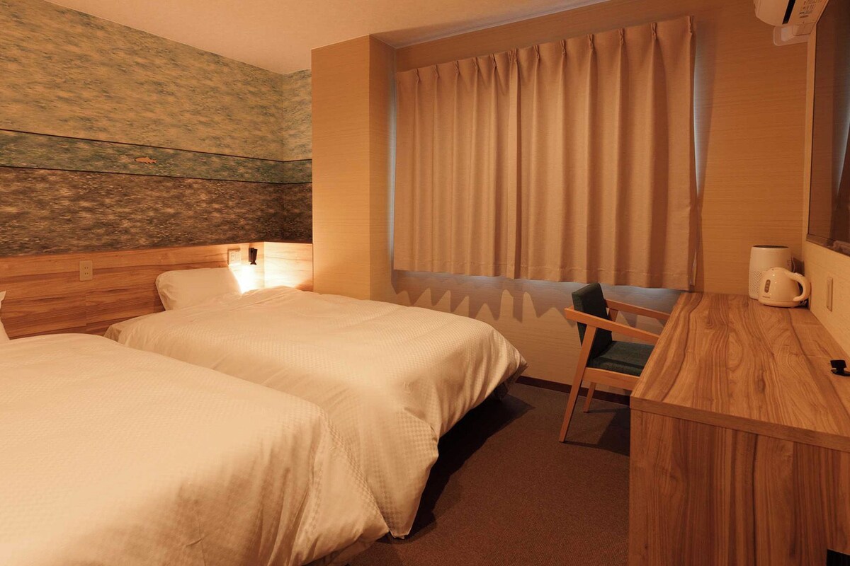 Habitación de hotel con dos camas, un escritorio de madera, una silla, un ventanal con cortinas y una pared con textura.