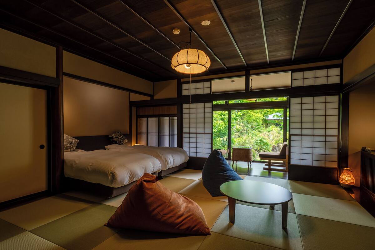 Un dormitorio tradicional japonés con suelo de tatami, una cama, una mesita y vistas a la vegetación exterior.