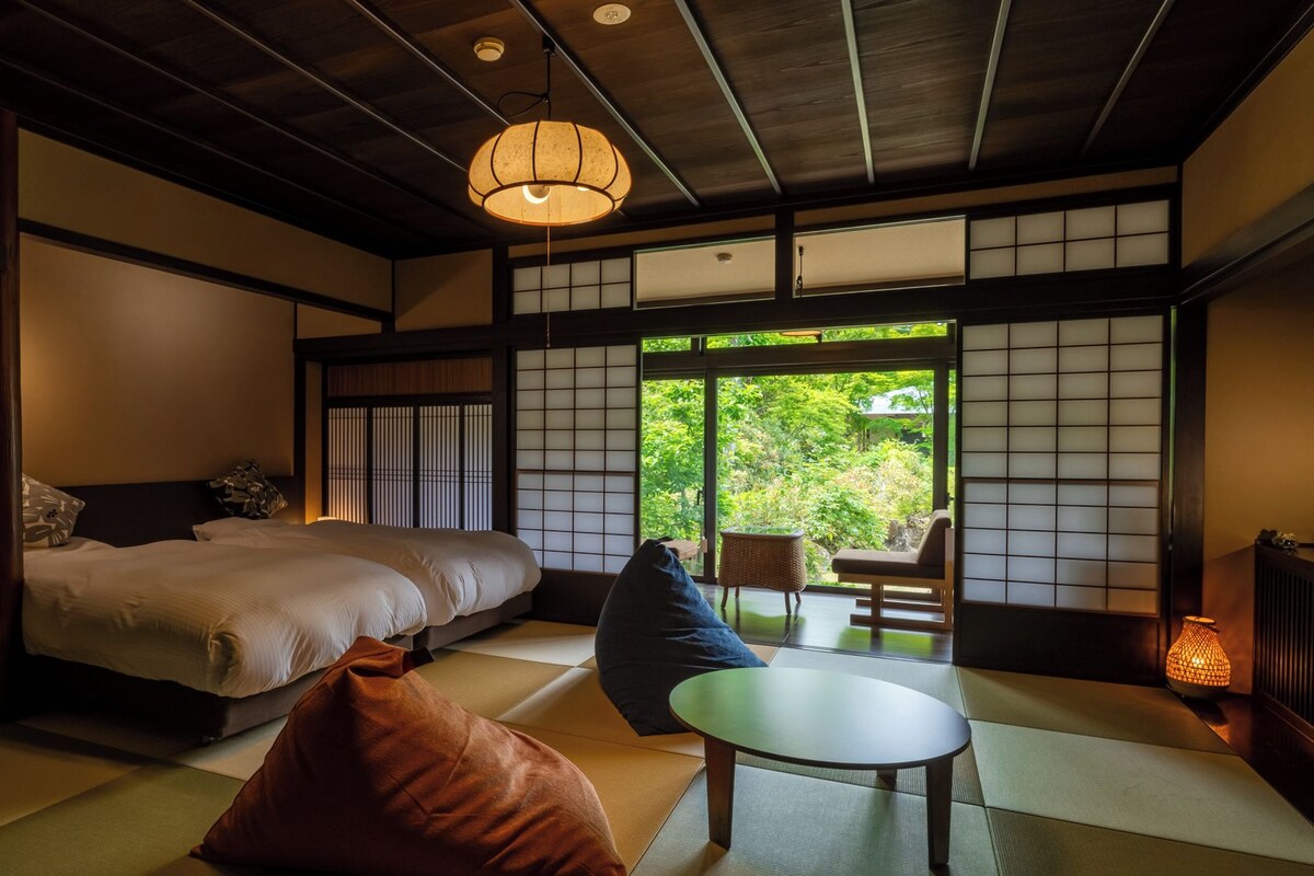 A traditional Japanese-style room with tatami flooring, a bed, a table, and a view of greenery outside.