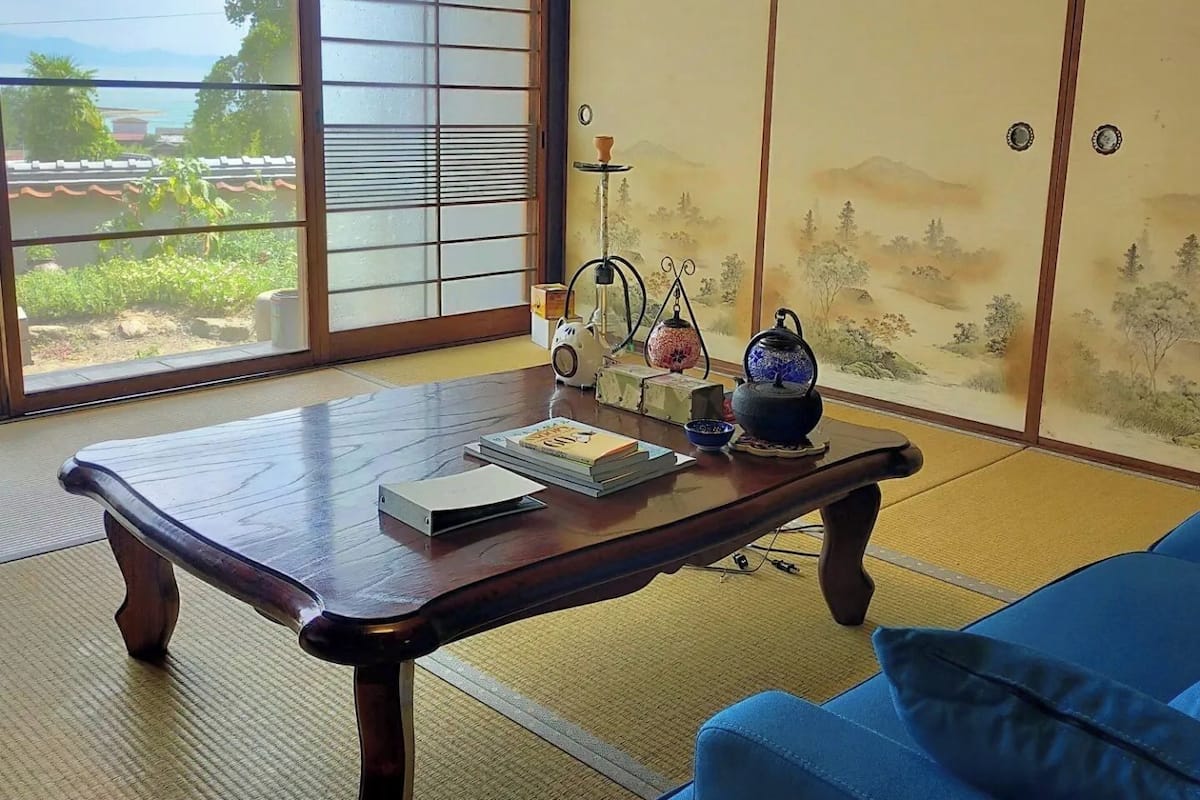 Una habitación con suelo de tatami, una mesa de madera con libros, un sofá azul y vistas a la vegetación a través de un shoji.