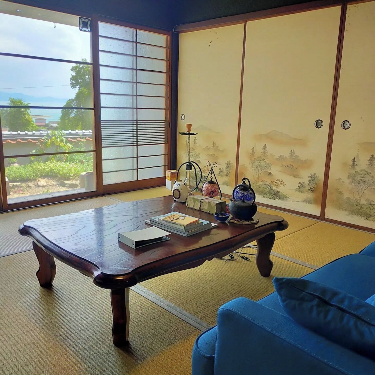Ein Zimmer mit Tatami-Boden, einem Holztisch mit Büchern, einem blauen Sofa und Blick ins Grüne durch einen Shoji-Bildschirm.