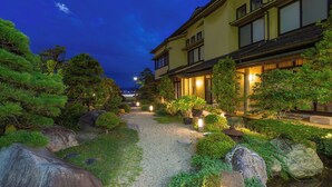 Un edificio de estilo japonés tradicional con un camino de grava, rodeado de follaje cuidado y rocas.
