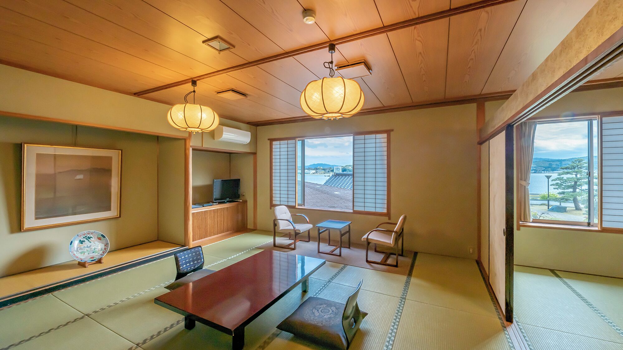 Um quarto japonês tradicional com tatames, uma mesa baixa, cadeiras, uma televisão e janelas grandes com biombos shoji.