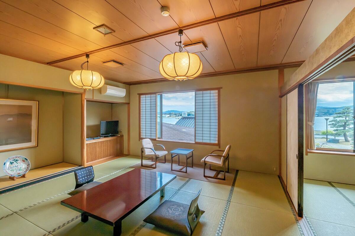 A traditional Japanese room with tatami flooring, a low table, chairs, a television, and large windows with shoji screens.
