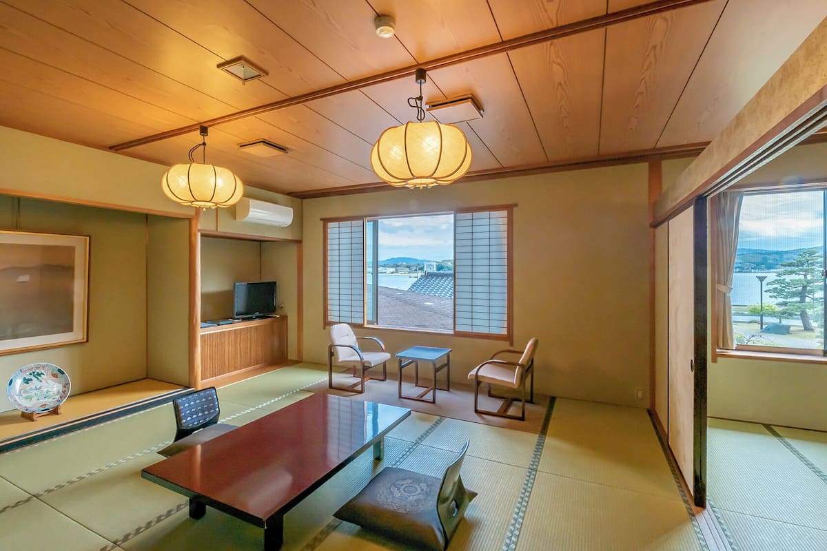 Una habitación japonesa tradicional con suelo de tatami, una mesa baja, sillas, un televisor y ventanas amplias con paneles shoji.