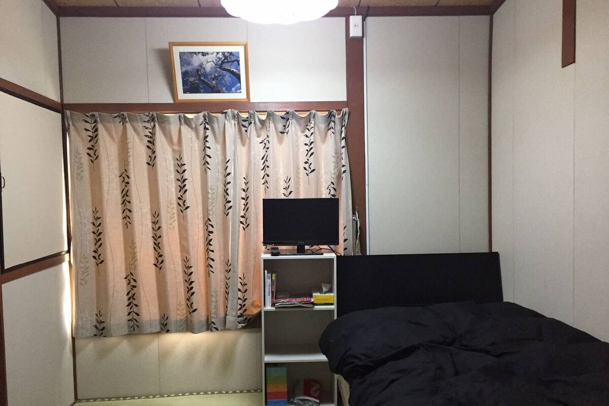 Un dormitorio con cama, televisor, estantería y ventana con cortinas.