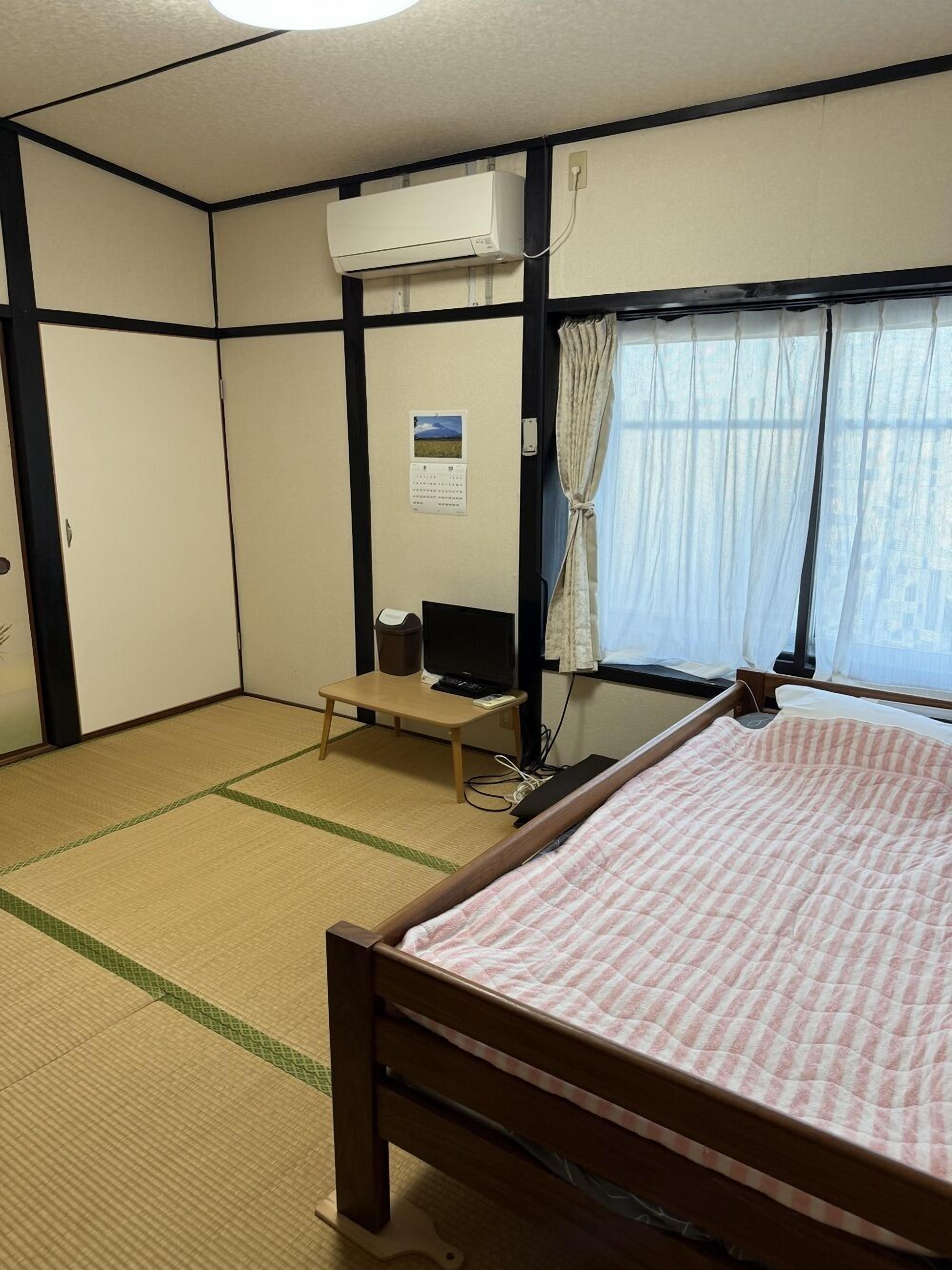 Una habitación con suelo de tatami, una cama con colcha rosa, aire acondicionado, un escritorio con ordenador, un calendario y una ventana con cortinas.