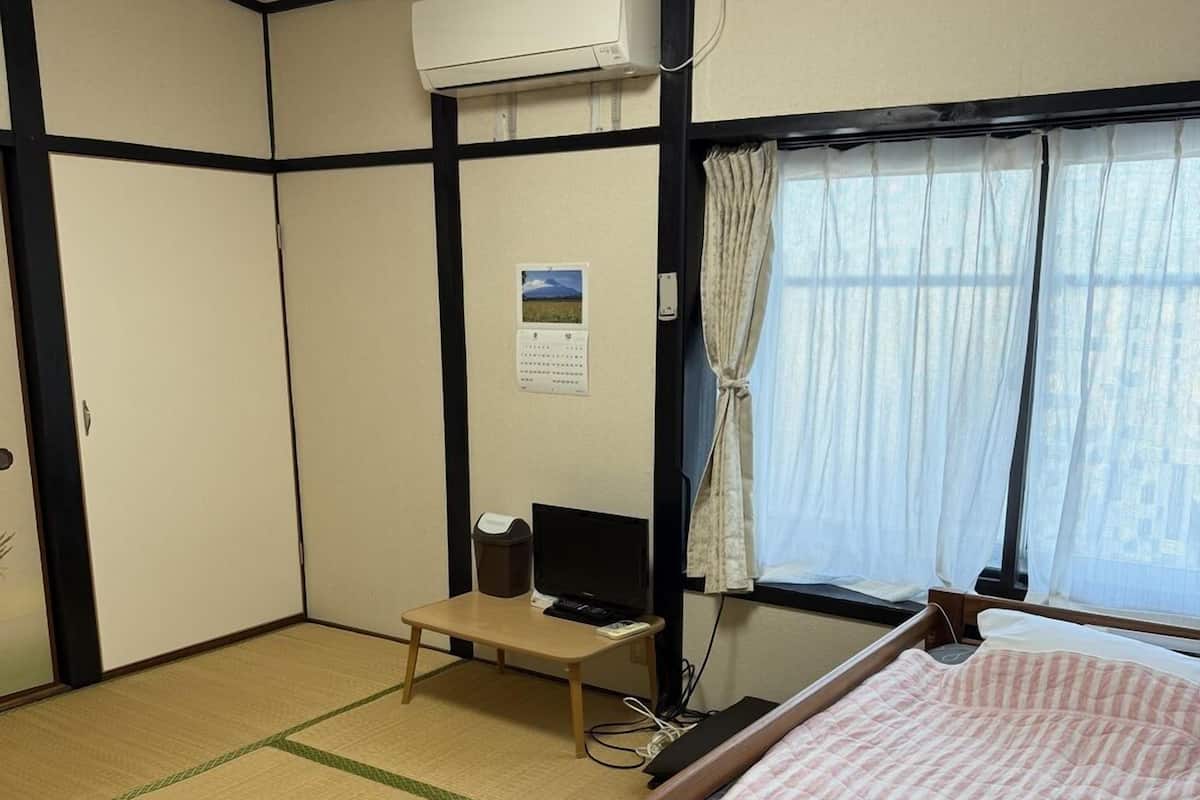 Una habitación con suelo de tatami, una cama con colcha rosa, aire acondicionado, un escritorio con ordenador, un calendario y una ventana con cortinas.