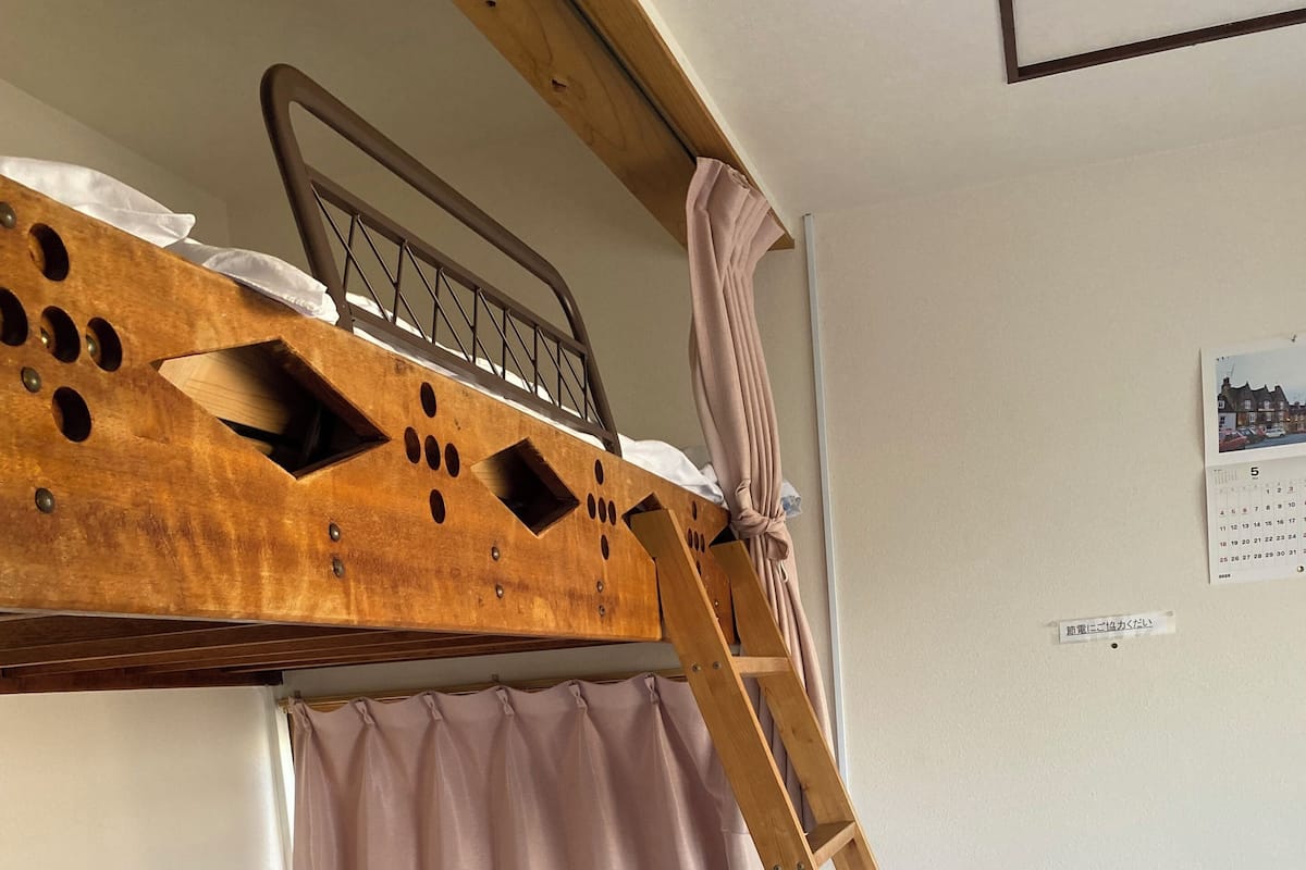 Una cama individual con escalera, una cama de solte con una manta estampada, una mesita con superficie de vidrio y un calendario en la pared.