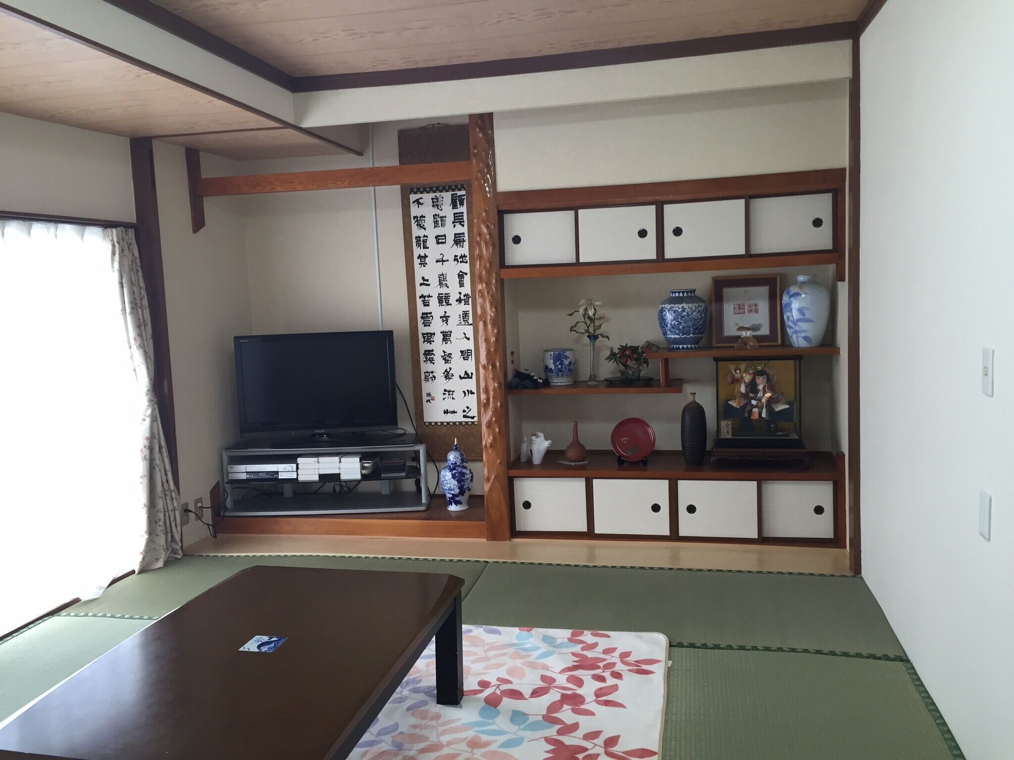 Una habitación japonesa tradicional con suelo de tatami, televisor, aparador con adornos y un shoji con caligrafía.