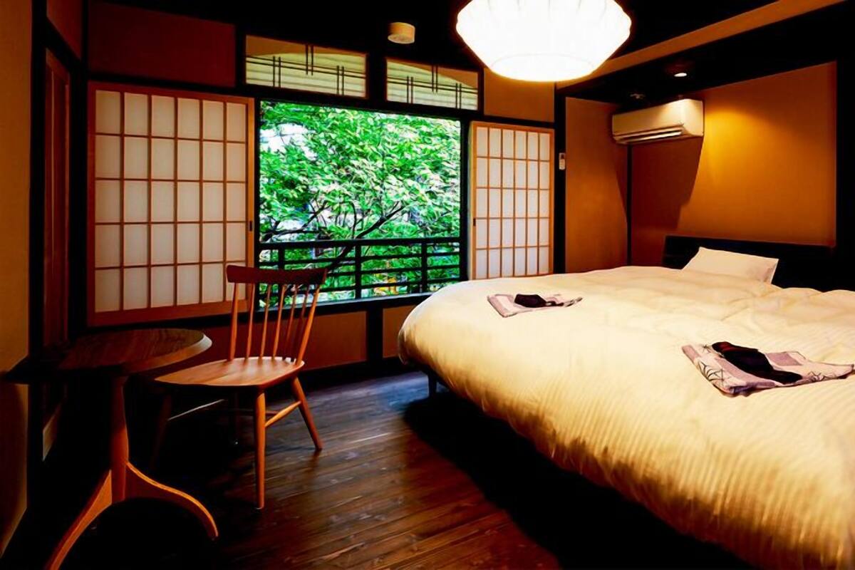 Habitación de hotel con una cama grande, una mesita pequeña, una silla y vistas a zonas verdes a través de una ventana con paneles translúcidos tradicionales japoneses.