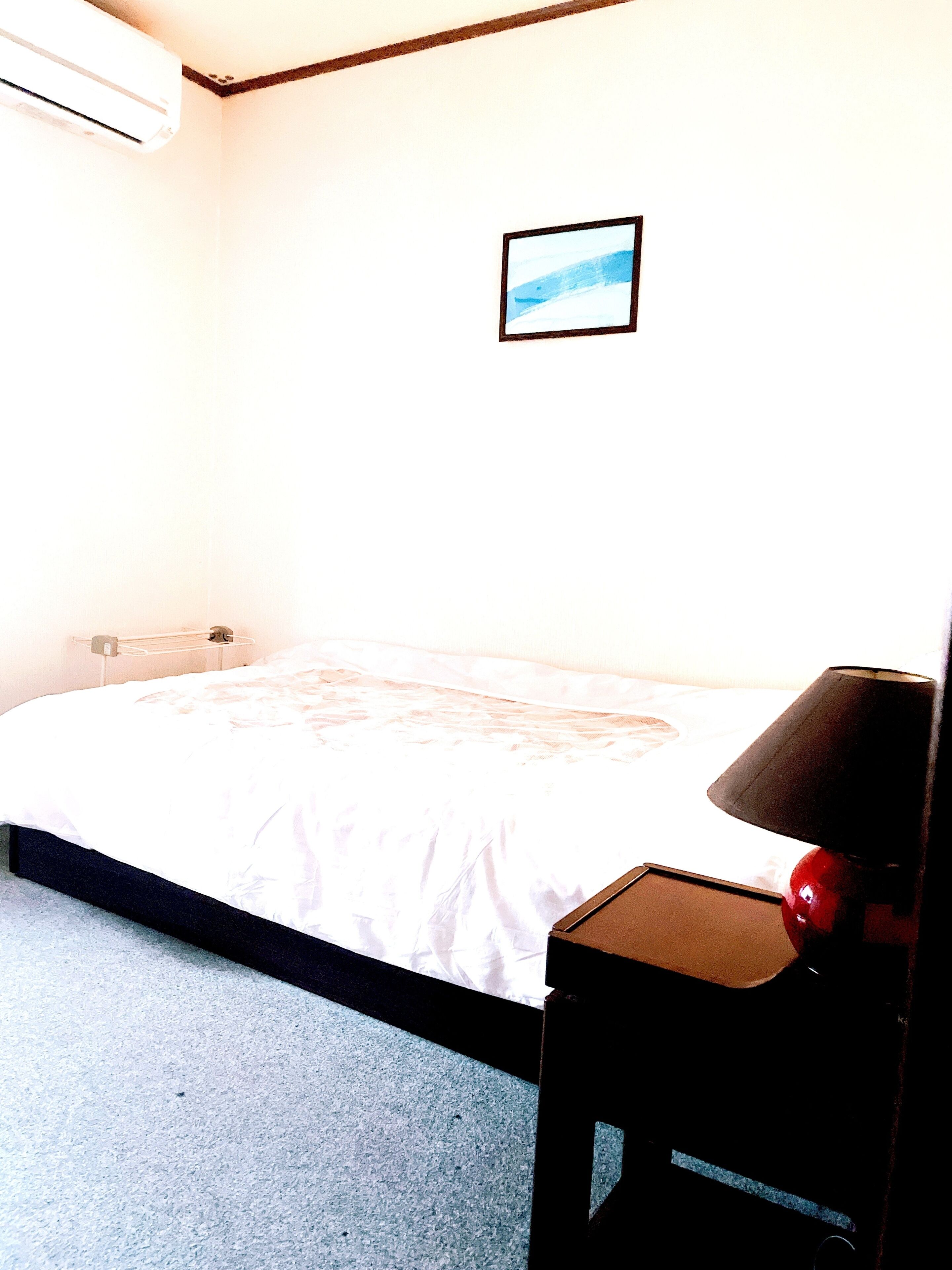A bedroom with a bed, a bedside table with a lamp, a wall-mounted air conditioner, and a framed picture on the wall.