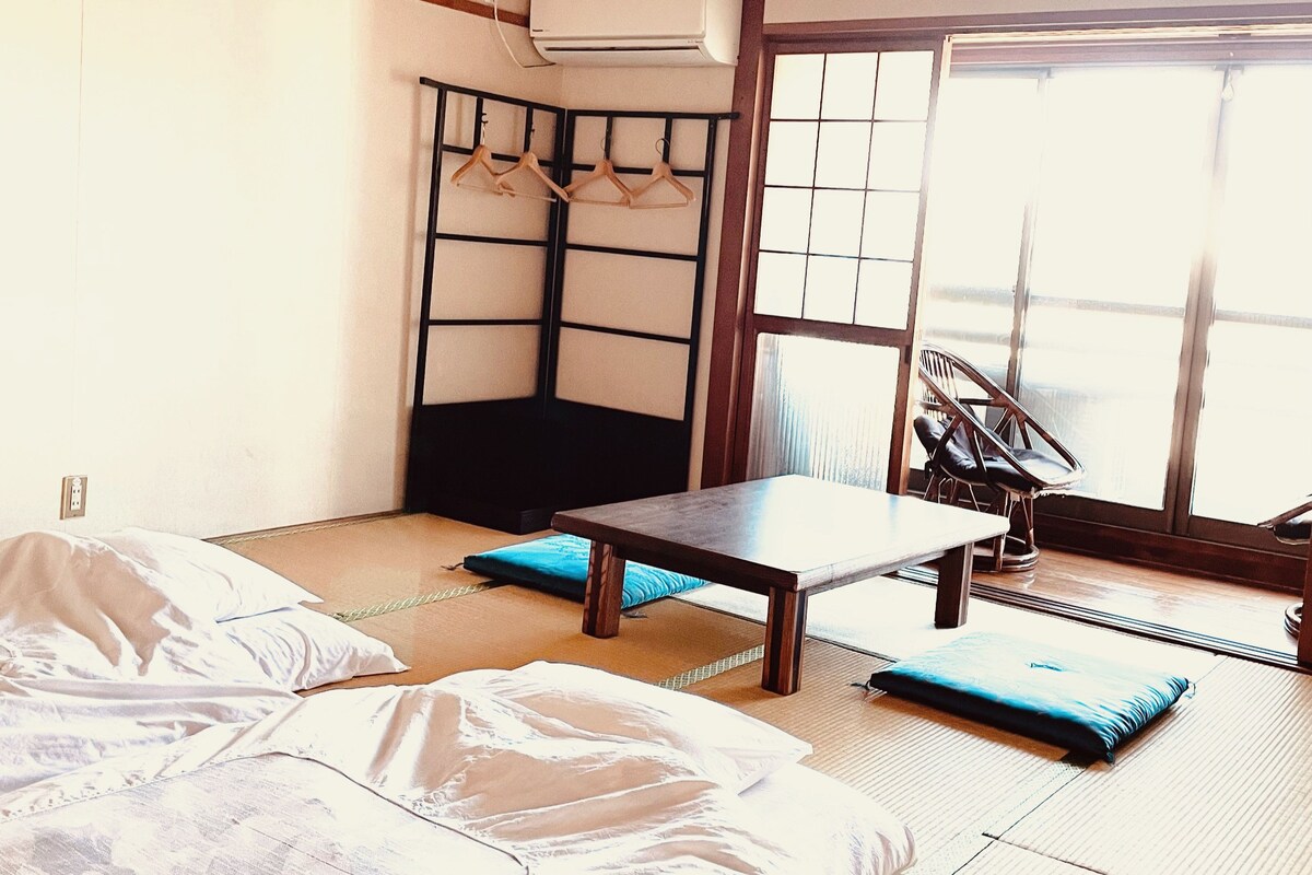 A room with tatami flooring, a futon bed, a low table, a chair, a hanging rack, and a sliding door.