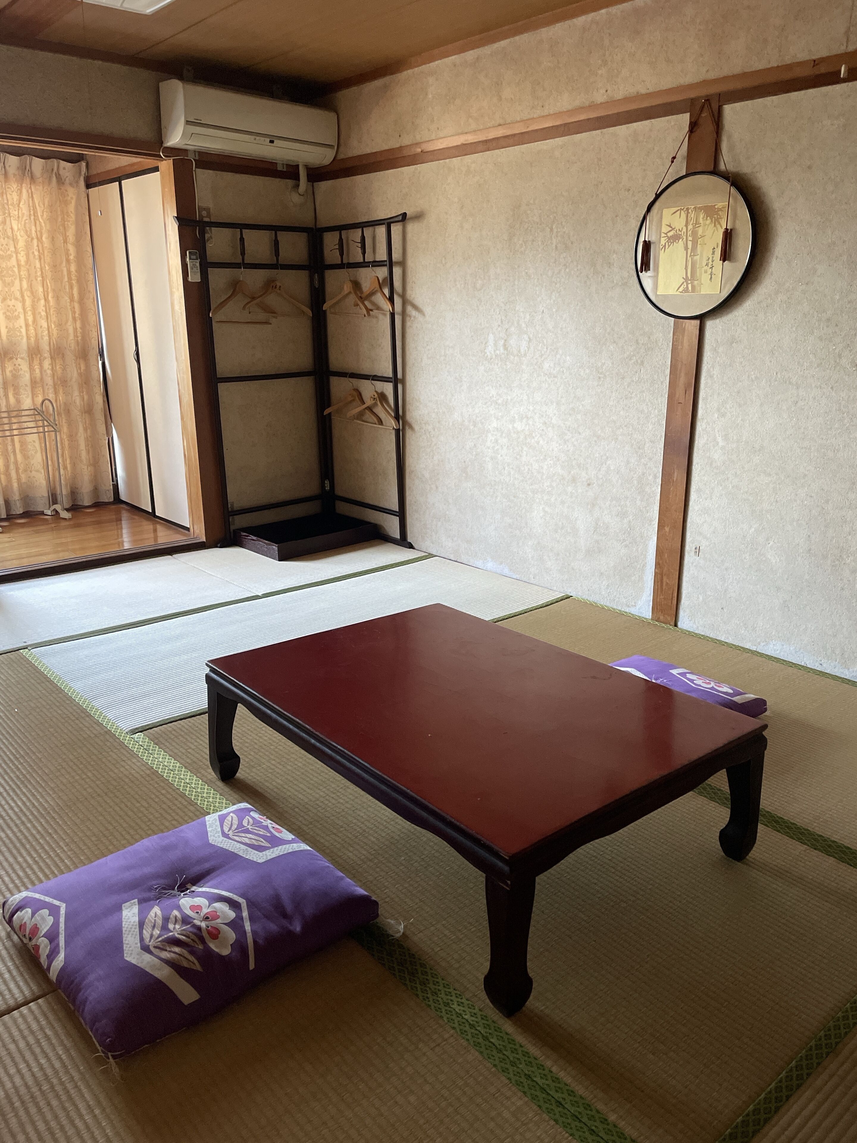 Une pièce avec un sol recouvert de tatamis, une petite table basse, un coussin, un miroir suspendu et une armoire.