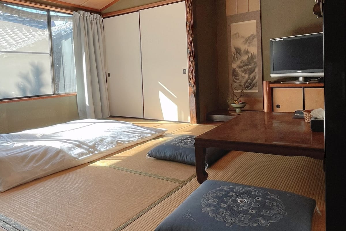 A traditional Japanese-style room with tatami flooring, a bed with white bedding, a television, and a sliding door.