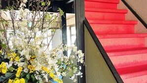 A staircase with red steps, a blue carpeted floor, and a large vase with white and yellow flowers. - Monzenya (Tokoname)