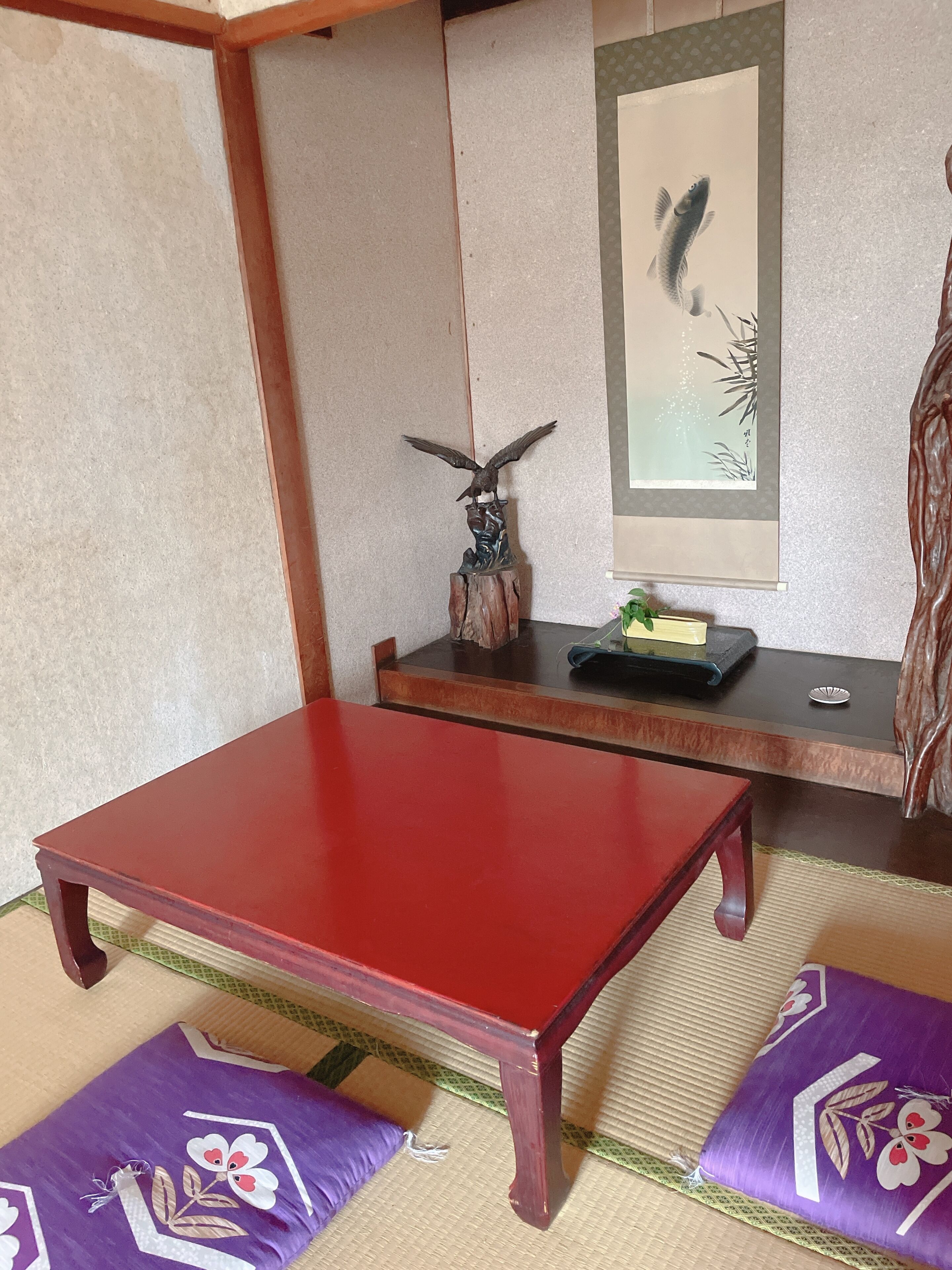 A traditional Japanese room with tatami flooring, a red table, purple cushions, and a wall with a framed artwork.
