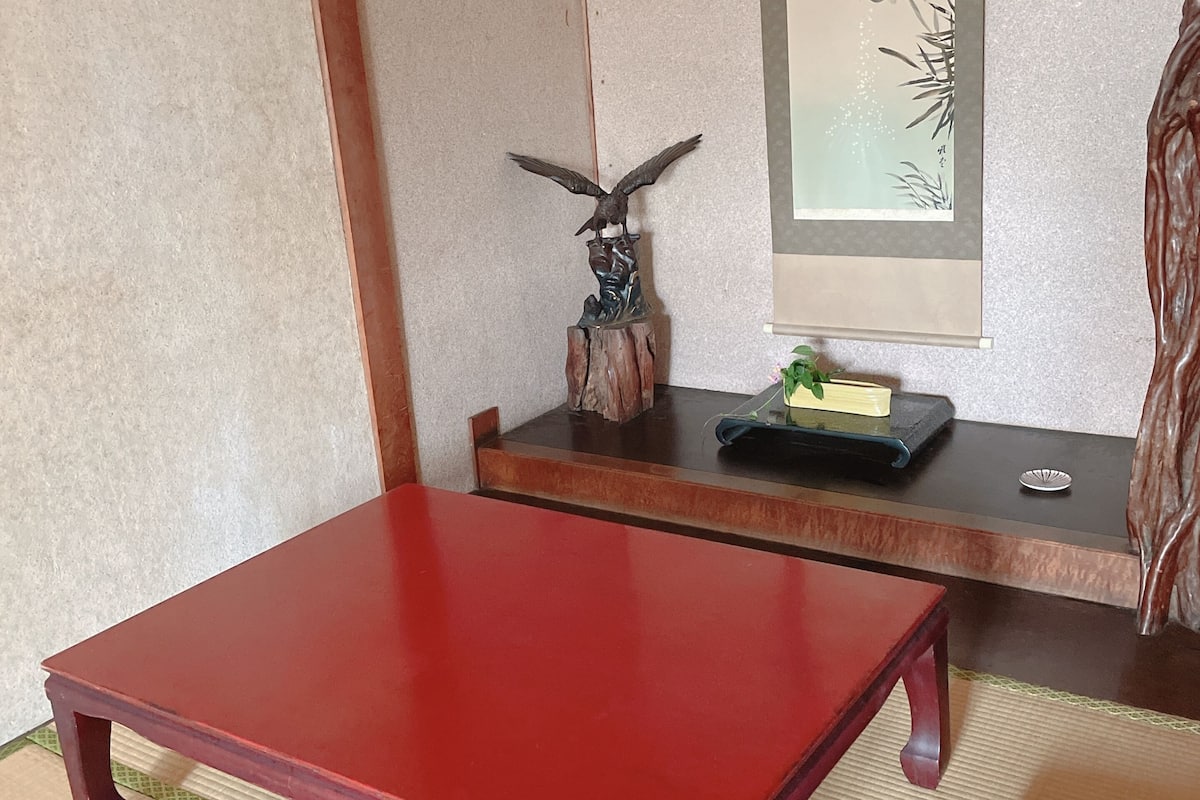 A traditional Japanese room with tatami flooring, a red table, purple cushions, and a wall with a framed artwork.