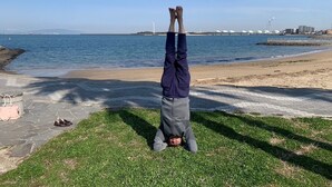 A person performing a handstand on grass near a beach with a clear sky and distant structures. - Monzenya (Tokoname)