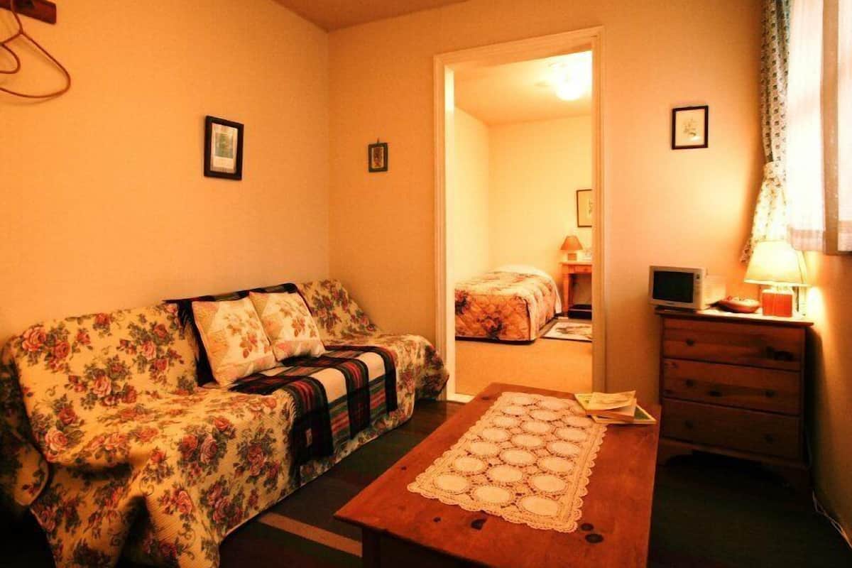 Ein Zimmer mit einem blumigen Sofa, einem Holztisch mit einer Spitzen Tischläufer, einer kleinen Holzkommode und einem Bett mit floralem Überwurf.