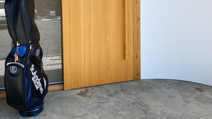 Uma porta de madeira com uma fechadura e uma pequena tranca eletrônica, uma bolsa preta e azul com a inscrição 'St. Andrews' e uma parede com caracteres japoneses e um desenho circular verde.
