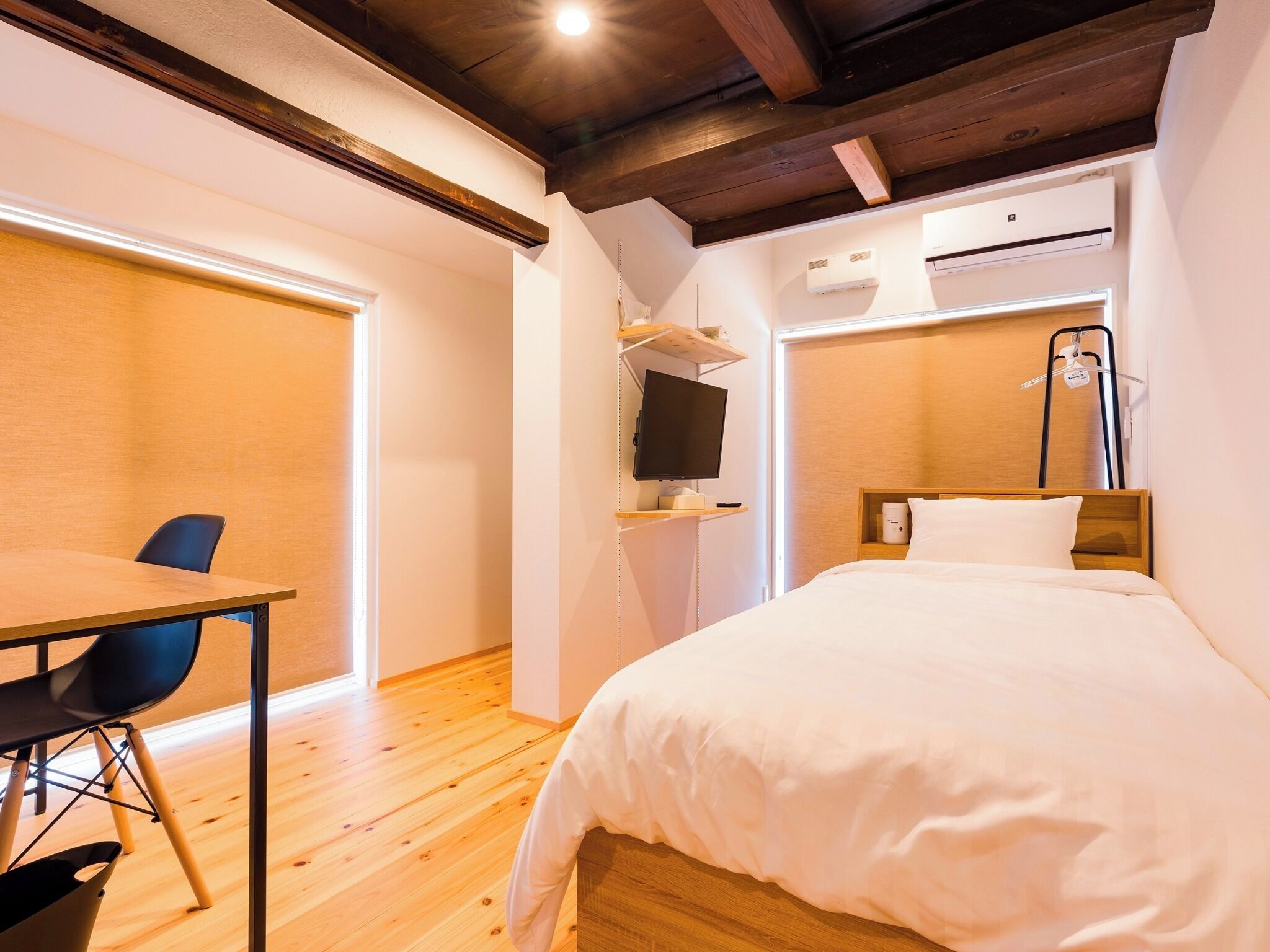 Ein Hotelzimmer mit Holzboden, einem Bett mit weißer Bettwäsche, einem Schreibtisch mit Stuhl, einem Fernseher und einer Klimaanlage.