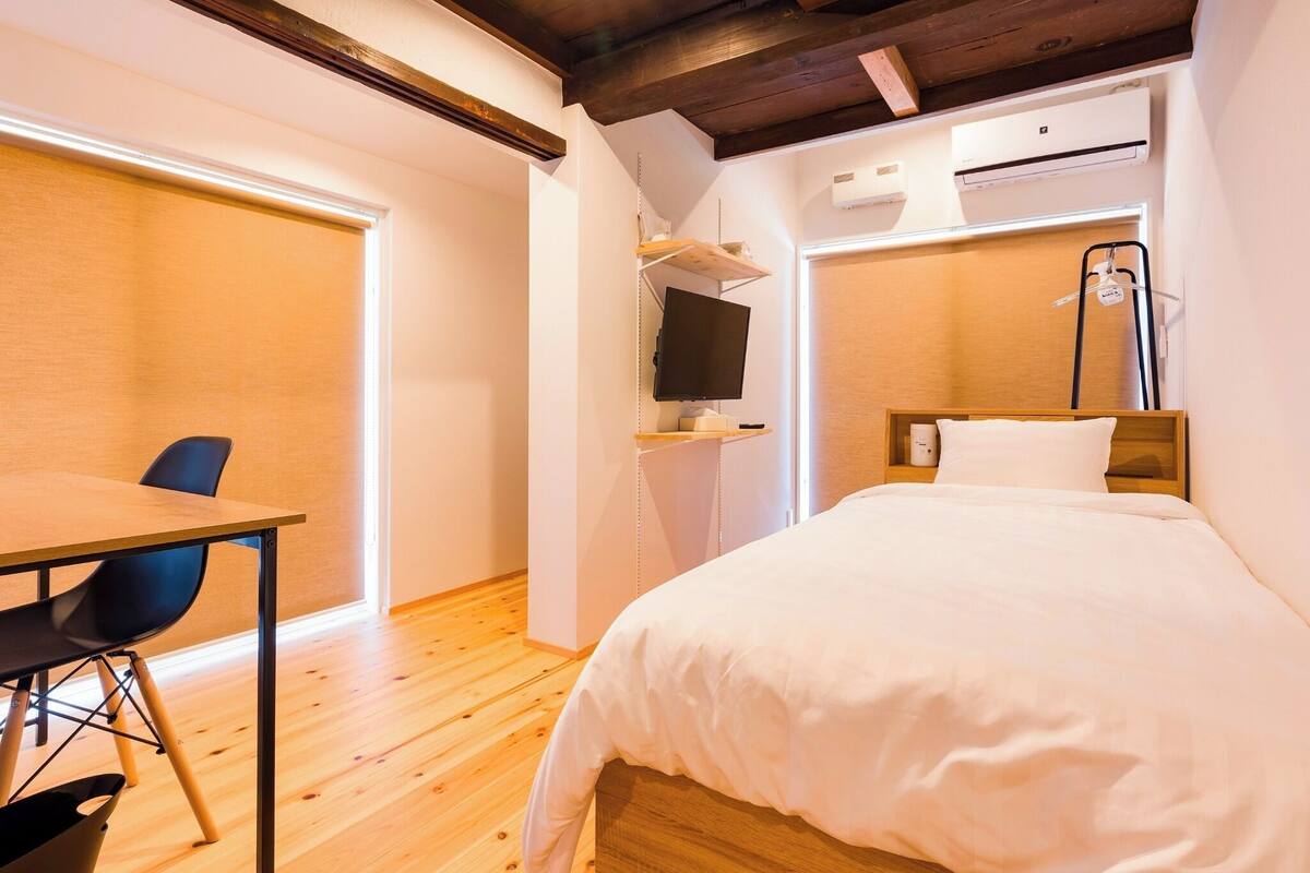 A hotel room with a wooden floor, a bed with white bedding, a desk with a chair, a television, and an air conditioning unit.