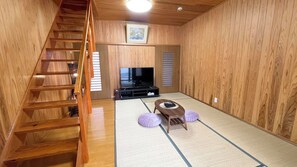 A room with a wooden staircase, a flat-screen TV, tatami mats, and a low wooden table with cushions. - Matsue no Sato (Matsue)