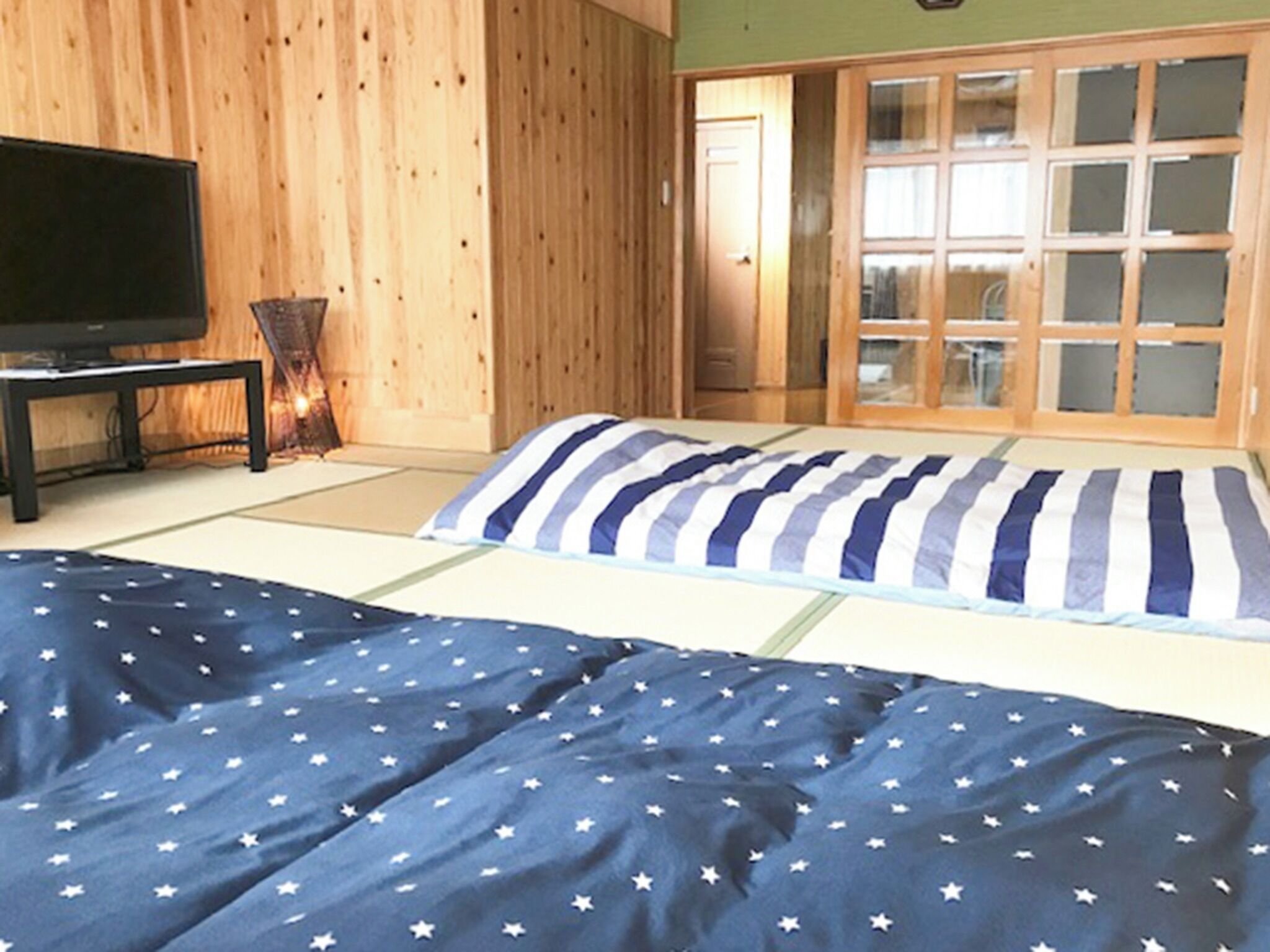 Ein Zimmer mit Tatami-Boden, einem Bett mit blau-weiß gestreifter Decke, einem Fernseher auf einem Schrank und einer Schiebetür, die zu einer Küchenzeile führt.