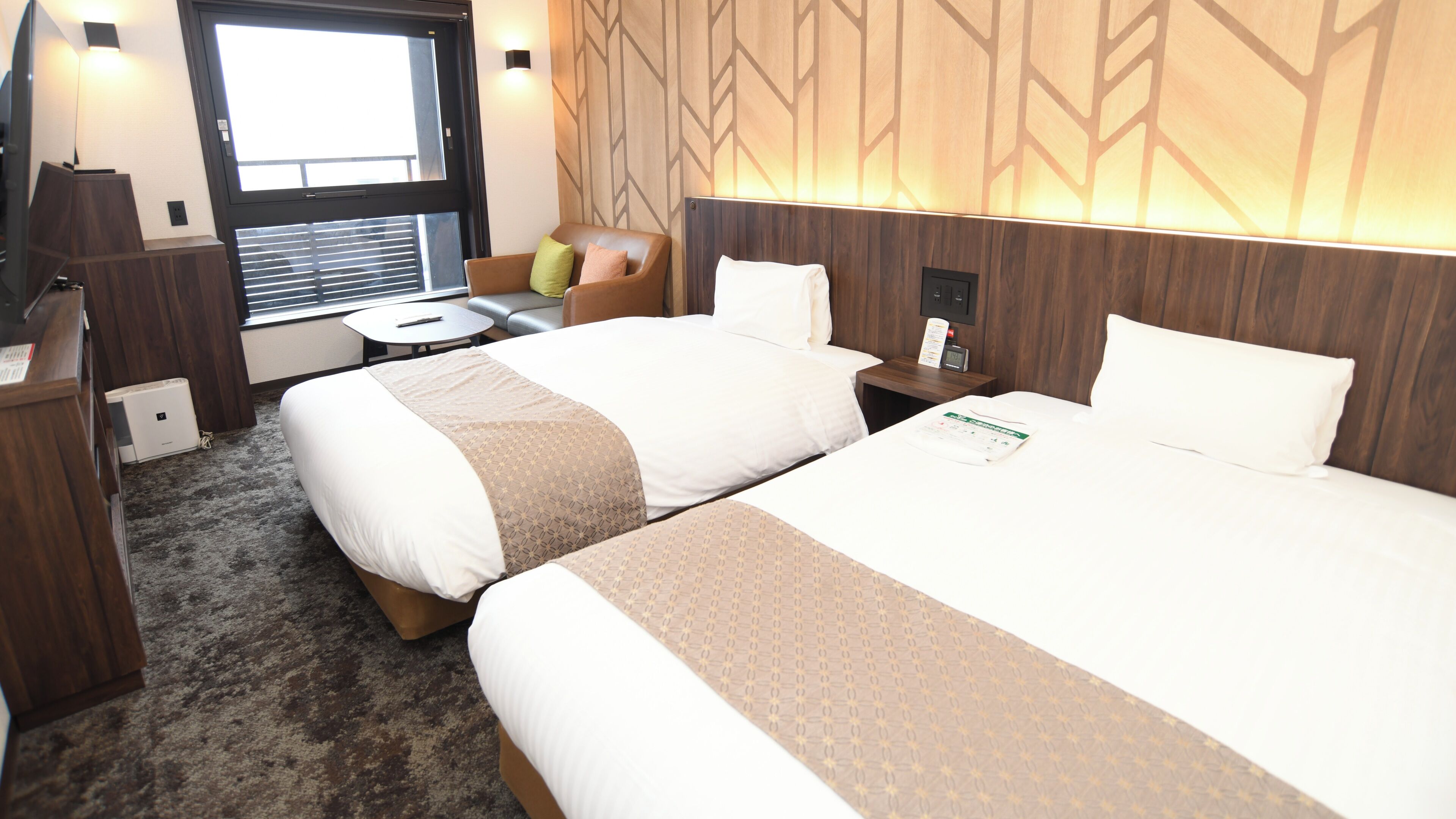 Quarto de hotel com duas camas, uma escrivaninha, uma cadeira, uma mesinha e uma janela com persianas.