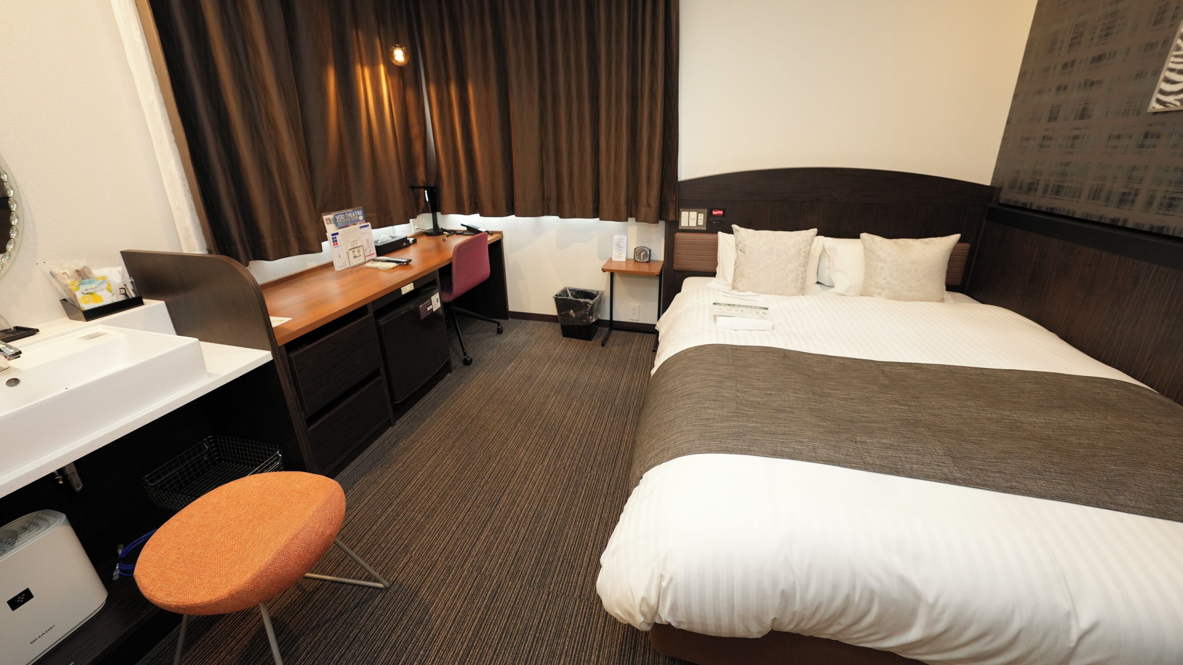 Quarto de hotel com cama, escrivaninha com cadeira, mesa pequena com abajur, lixeira e janela com cortinas.