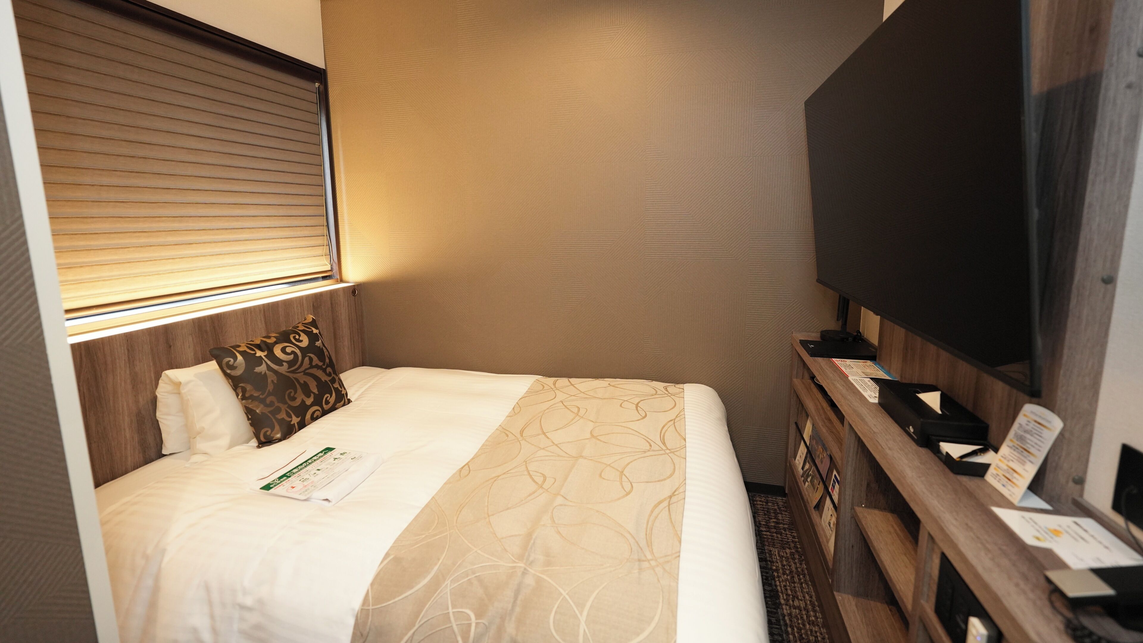 Quarto de hotel com cama, televisão, secretária de madeira e janela com persianas.