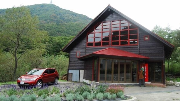 A two-story wooden building with large windows and a red roof, surrounded by greenery and a parked car. - Tairappyouchaya (Yuzawa)